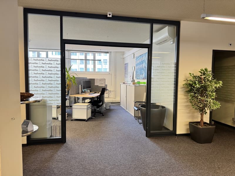 Office space with glass walls, desk, chairs, air conditioning, plants, and a trash bin.