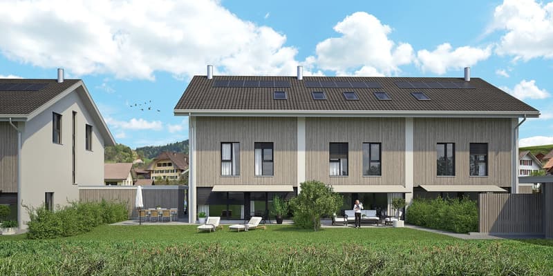 Two-story modern house with gray siding, large windows, and a balcony. The house has a patio area with outdoor furniture and is surrounded by a grassy lawn. Solar panels are visible on the roof.