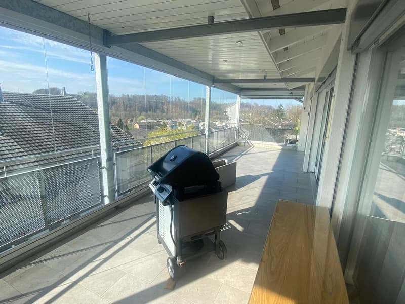 Balcony with tiled floor, glass railings, a wooden bench, a large window view of the city.