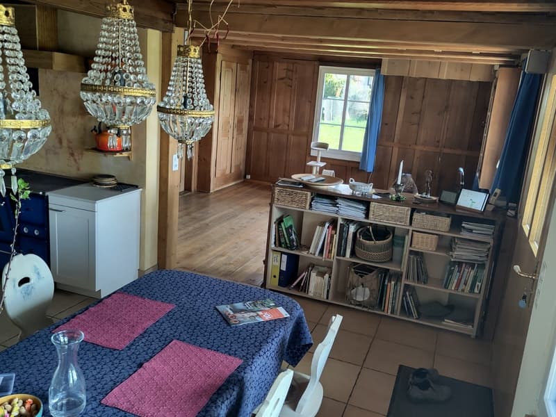 dining room with wooden floor, wooden beams, chandeliers, table, chairs, shelf with books and baskets, kitchen area with stove and refrigerator