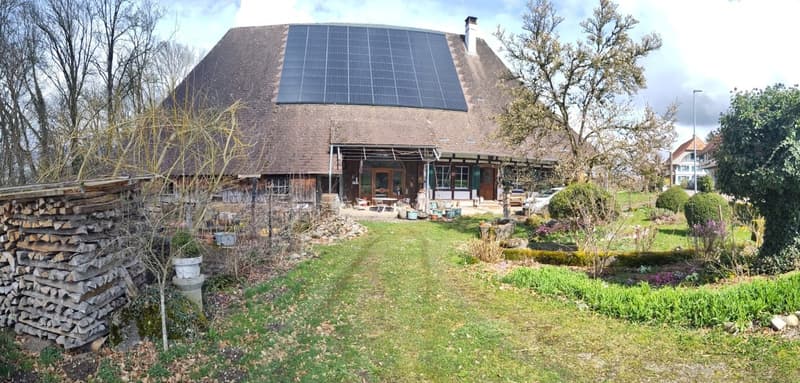Two-story brick house with a large solar panel array on the roof, surrounded by a well-maintained garden and yard with stacked firewood. The house has a rustic, eco-friendly appearance.