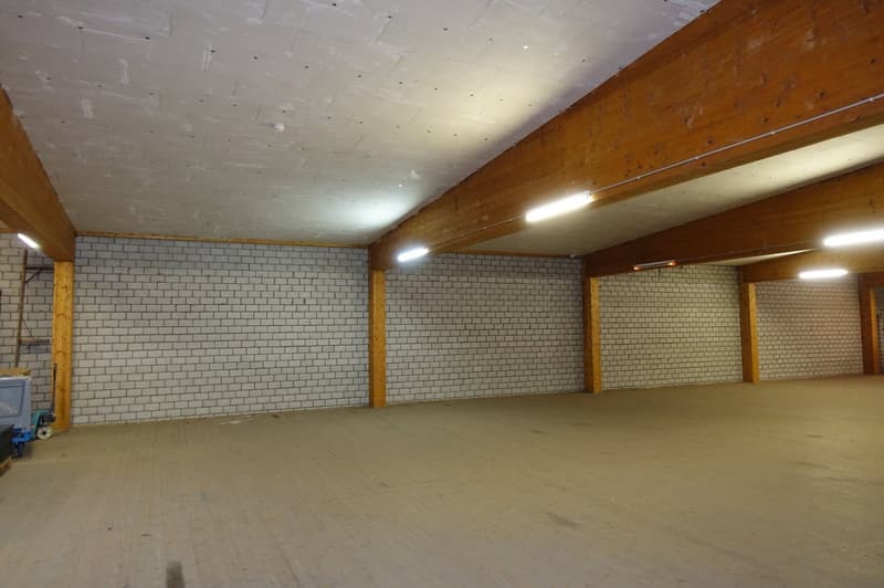 Large open space with concrete floor, white brick walls, and exposed wooden beams and ceiling. The space appears to be a warehouse or storage facility with overhead lighting.