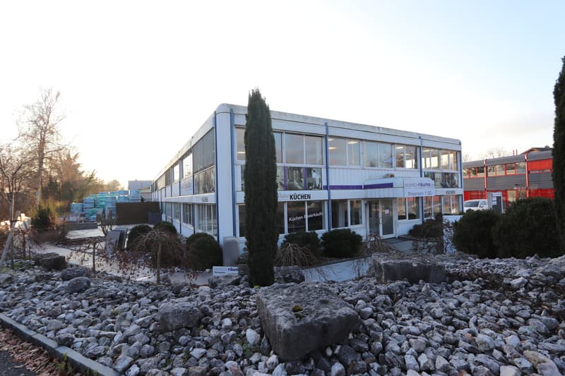 Modern office building, large glass windows, white exterior, modern design, landscaped garden with large rocks, visible signage for kitchen sales