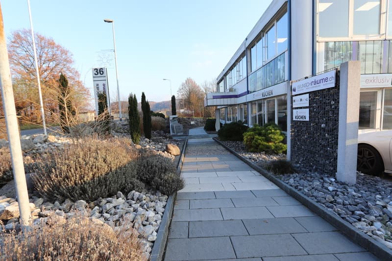 Modern building with glass facade, entrance area with sidewalk, landscaping with shrubs and gravel.