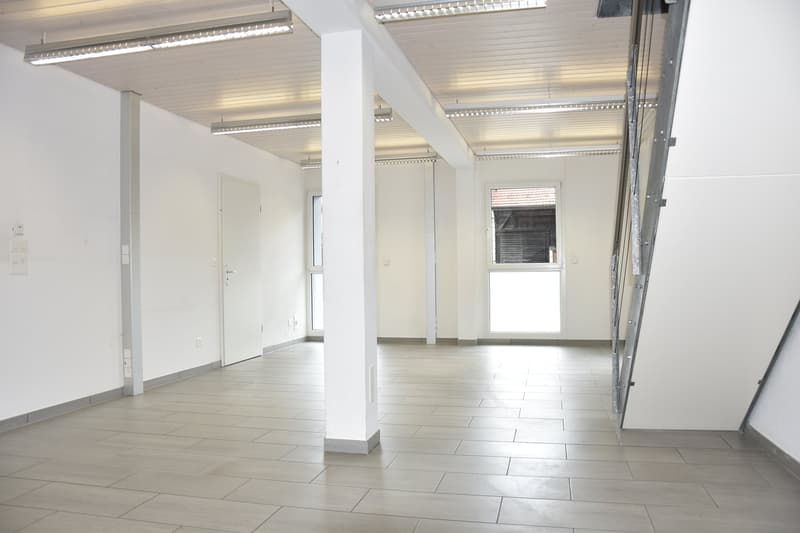 A large, empty room with tiled flooring and white walls. There are three doorways and two large windows in the room. Four white columns are visible in the room.