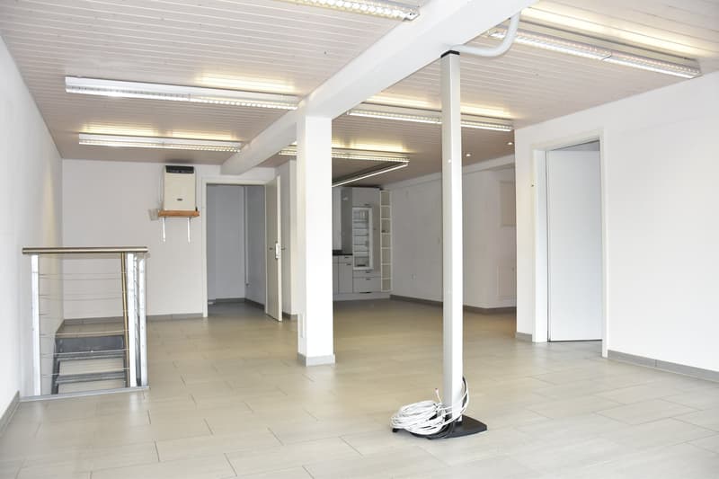 Spacious basement with tiled floors, recessed ceiling lights, white walls, metal handrail, door leading to the entrance