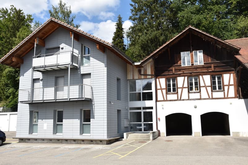Modern building, 2 levels, white exterior, brown roof, glass windows, balconies on the second floor, garage entrance, parking area