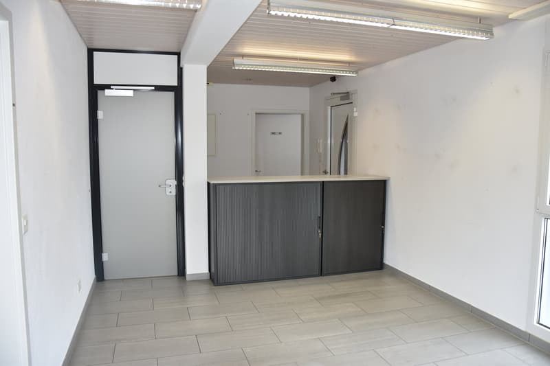 Empty room with tiled floor, double doors on left, double doors and cabinets in middle, two large windows on the right, a picture hanging, fluorescent lights on ceiling