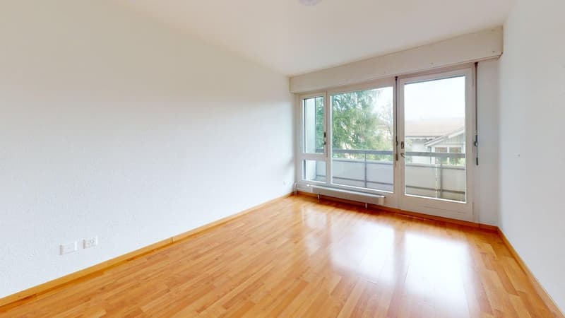 Empty room, hardwood floors, white walls, sliding doors leading to balcony, windows with view of trees and house, wall mounted light, power outlets on the wall.