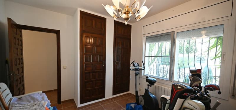 This image shows a room with wooden doors, a chandelier, and a window overlooking a balcony or terrace area. There is also an exercise bike visible in the room.
