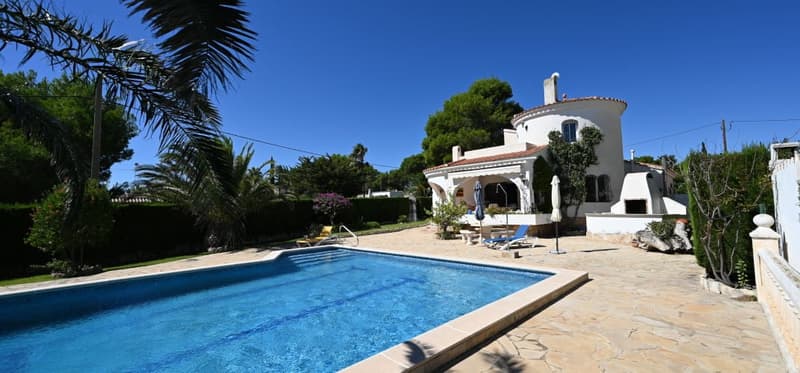 Two-story Mediterranean-style villa with a swimming pool, surrounded by lush greenery and palm trees. The villa has a balcony or terrace and a tiled roof.