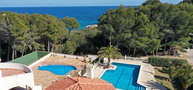 This image shows a scenic coastal property with a swimming pool and beautiful ocean views. The property has a large outdoor area with a patio, palm trees, and lush greenery surrounding the pool. The blue waters of the ocean can be seen in the background,