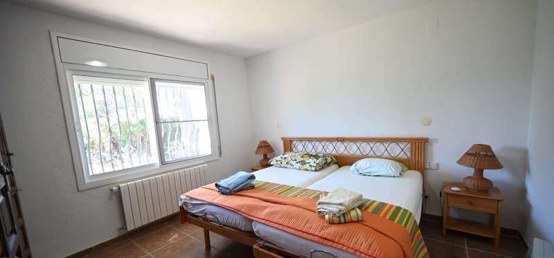 Spacious bedroom with large windows, wooden bed frame, bedside tables with lamps, and tiled flooring.