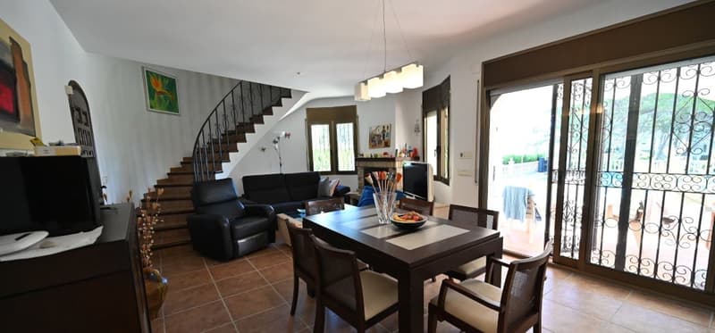 Open-concept living room and dining area, with a staircase leading to the upper level. The room features a large sofa, a dining table with chairs, and a sliding glass door leading to a balcony or terrace area. The decor includes artwork on the walls and a