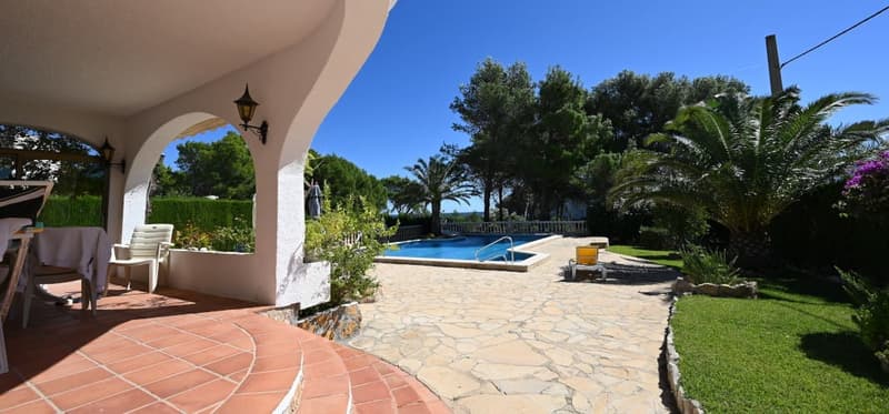 Two-story villa with a terrace, surrounded by a lush garden with palm trees and a swimming pool. The exterior features a stone patio and arched entryway.
