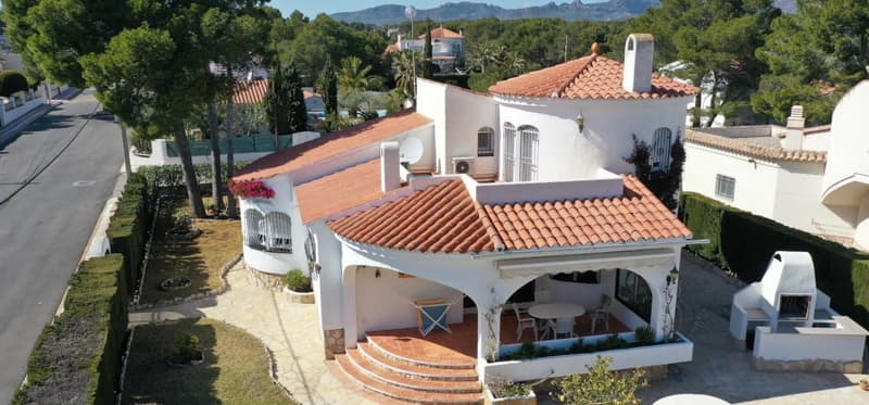 Two-story Mediterranean-style villa with red tile roof, white exterior walls, and arched windows. The house has a balcony or terrace, and there appears to be a parking area or driveway. The surrounding landscape includes palm trees and other greenery.