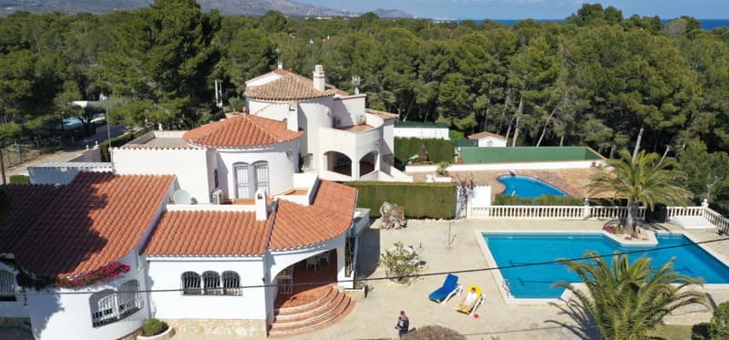 Large two-story Mediterranean-style villa with red tile roof, white exterior walls, and arched windows. The property has a swimming pool, balconies, and a garage. It is surrounded by lush greenery and palm trees, with a scenic view of the surrounding land