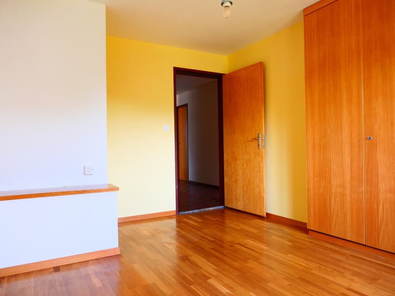The image shows a bedroom with yellow walls, wooden flooring, and a wooden wardrobe. There is a doorway leading to another room.