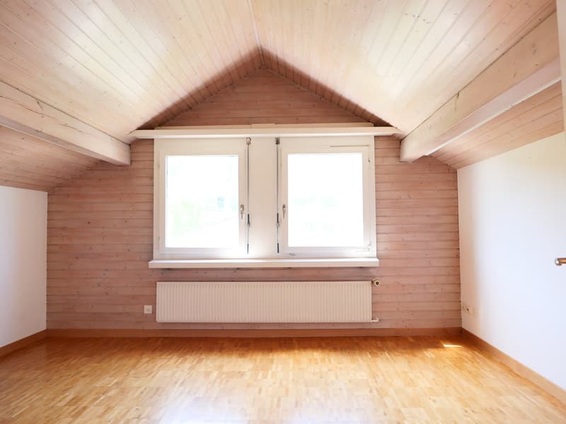 This image shows a spacious room with wooden walls and ceiling, a large window, and a radiator on the wall. The room has a hardwood floor and appears to be a living room or bedroom.