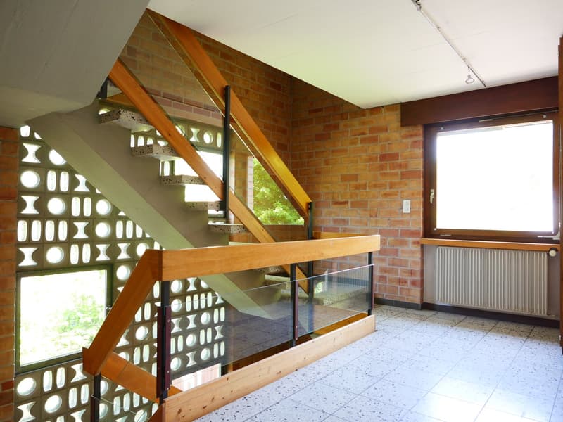 The image shows the interior of a building with a unique architectural design. The space features a staircase with a wooden railing and a glass partition, creating an open and airy feel. The walls are made of exposed brick, adding a rustic touch to the sp