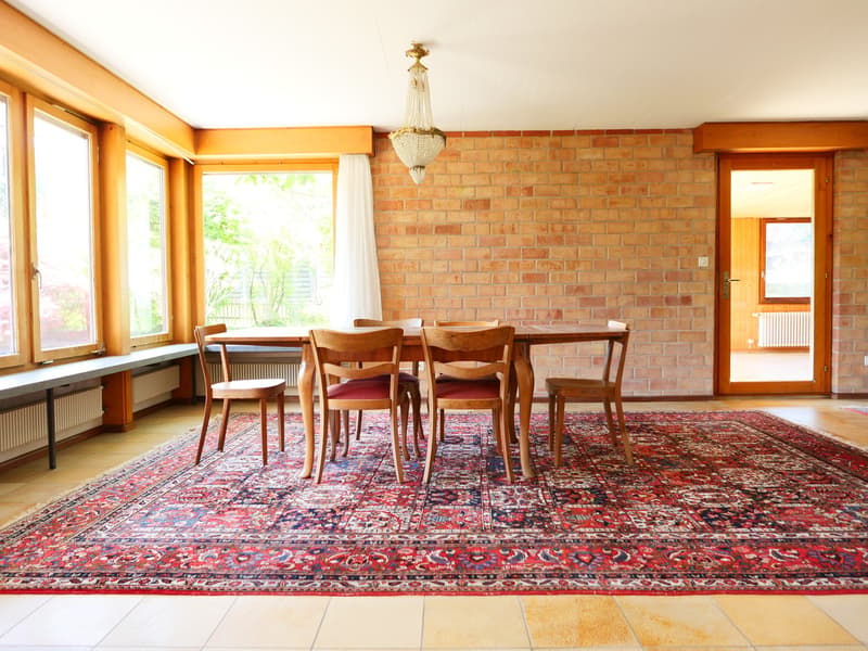 Spacious dining room with brick walls, large windows, wooden dining table and chairs, and a decorative chandelier. The room features a large, colorful patterned rug covering the floor.