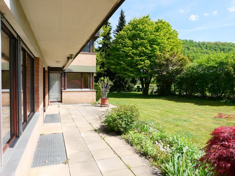 Two-story brick building with large windows, surrounded by a well-maintained garden with lush greenery, including trees and flowering plants. The building has a covered patio or balcony area overlooking the scenic outdoor space.