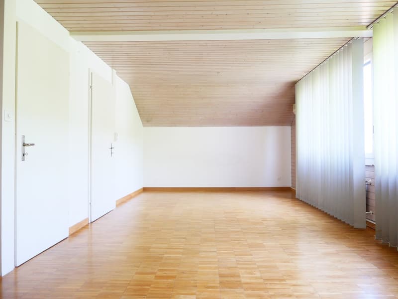 Large open room with wooden ceiling and floor, white walls, and a window with curtains. The room appears to be empty, with no furniture or decor visible.