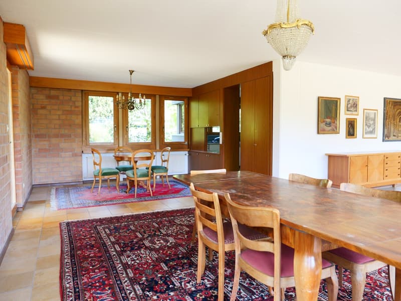 Spacious dining room with wooden furniture, including a large dining table and chairs. The room features a brick fireplace, hardwood floors, and large windows overlooking the outdoor scenery. The room is decorated with artwork and a chandelier, creating a