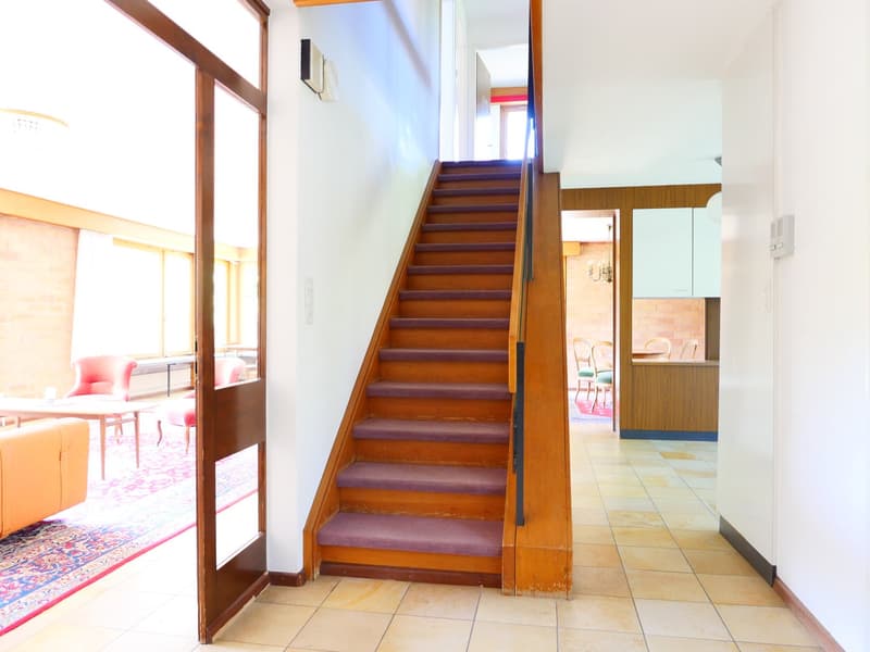The image shows the interior of a building, with a wooden staircase leading up to the second floor. The staircase has a railing and is surrounded by white walls. The floor is tiled, and there is a living room area visible through a doorway. The overall st