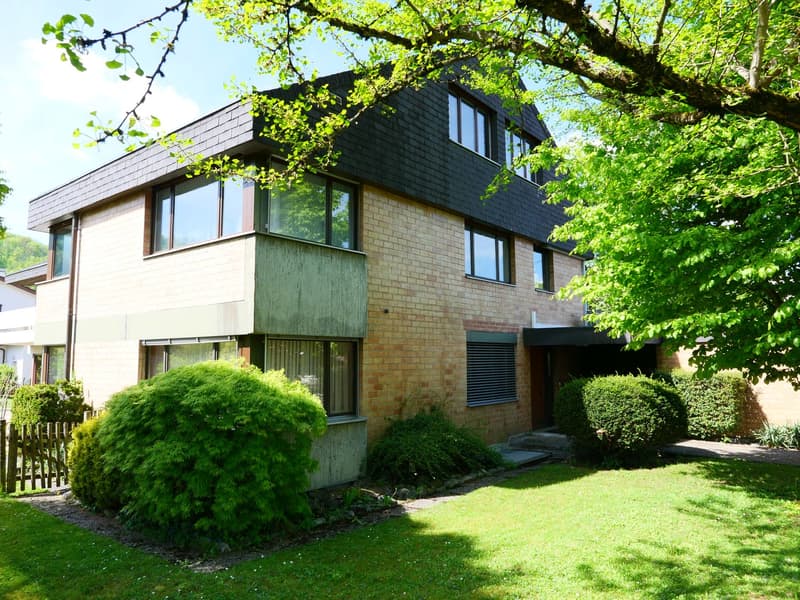 2-story house with brick and siding exterior, large windows, balcony, surrounded by lush greenery and trees