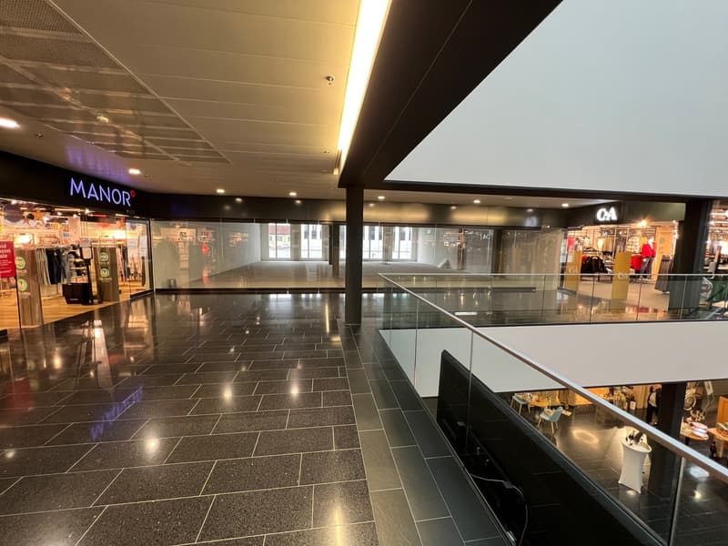 large shopping mall, empty, tiled floor, high ceiling, shops, sign for MANOR, light and glass railings