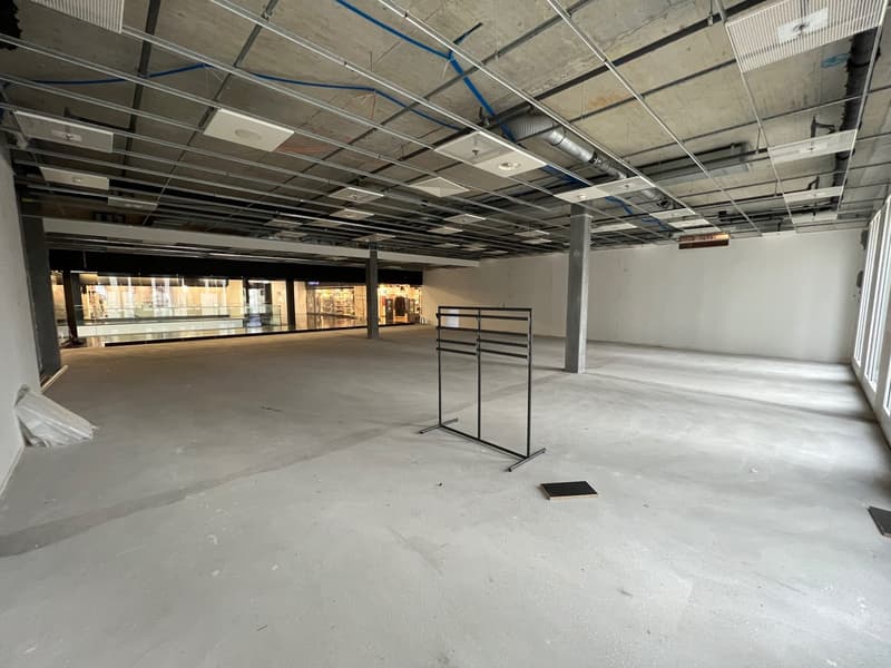 The image shows an interior of an empty building with a white concrete floor and exposed steel and metal beams with blue pipes attached.