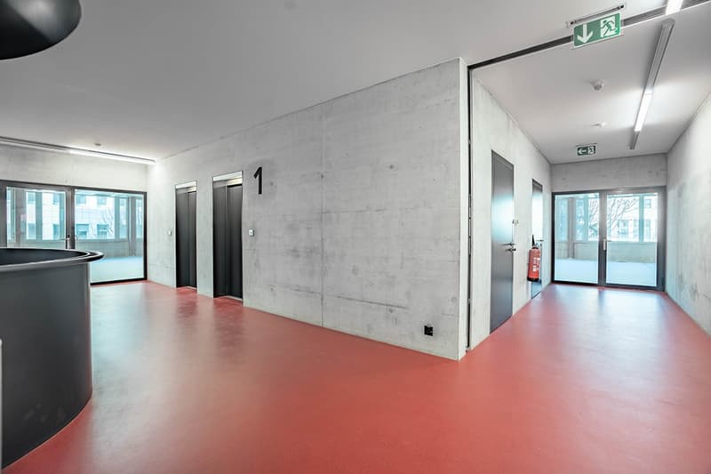 Concrete walls, red flooring, elevator, two entrances, glass door, fire extinguisher.