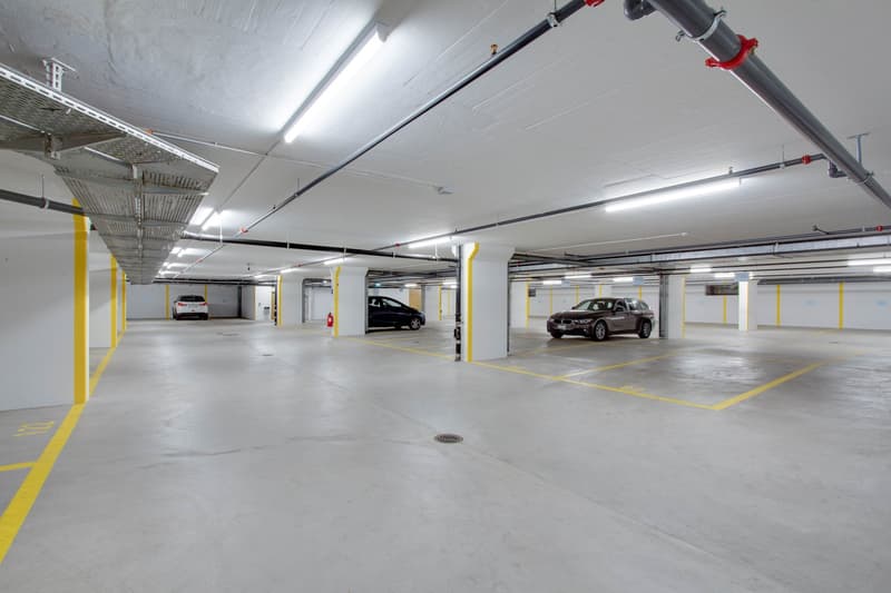 White walls, yellow and grey markings, fire extinguisher, fluorescent ceiling lights, parking spaces, 3 cars parked