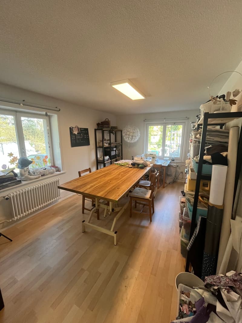 large room with wooden floor, long wooden table, chairs, shelves with various items, windows, radiator
