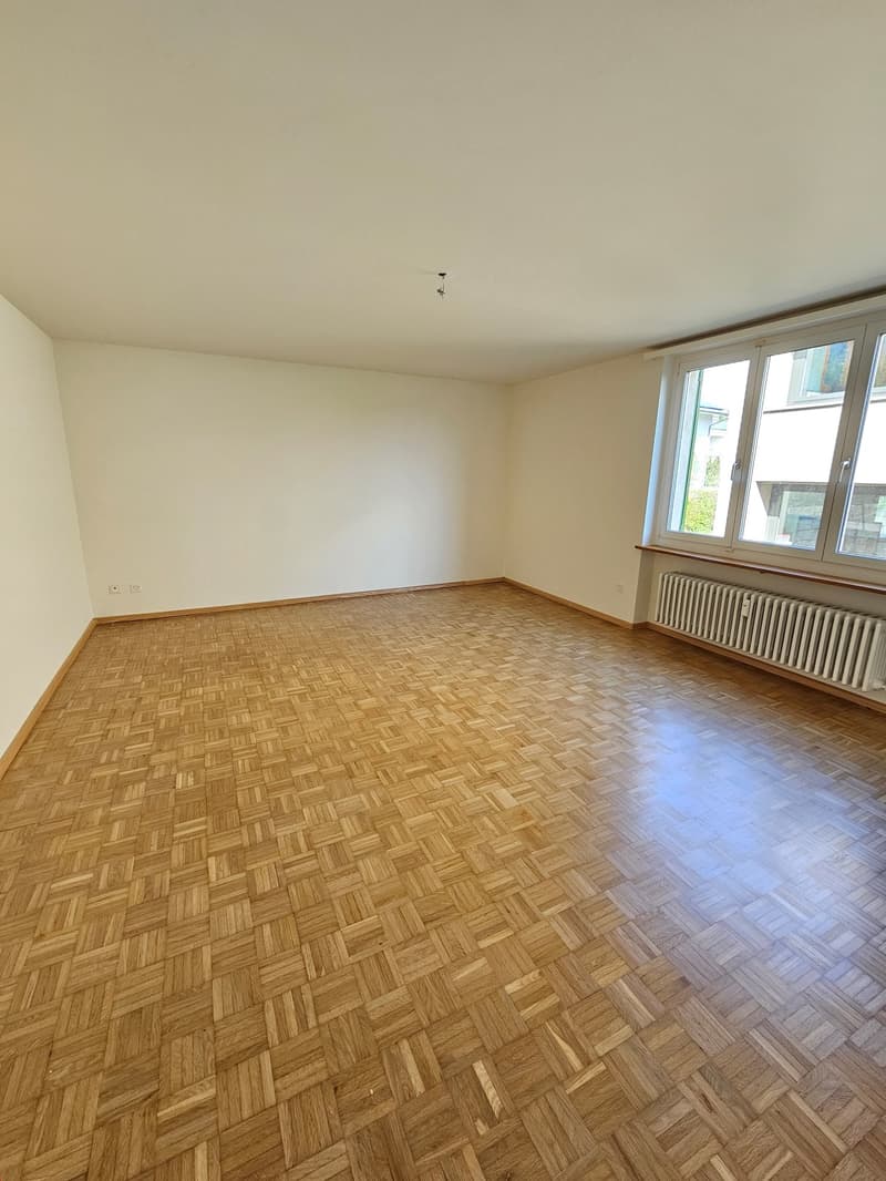 An empty room with a hardwood parquet floor, white walls, and a window that allows a view outside