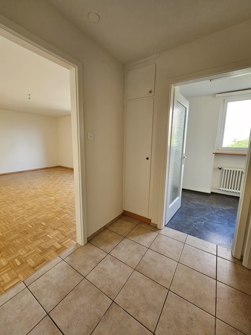 Empty room with wooden flooring and tiled flooring, white walls, and glass door leading to a bathroom with a white radiator.