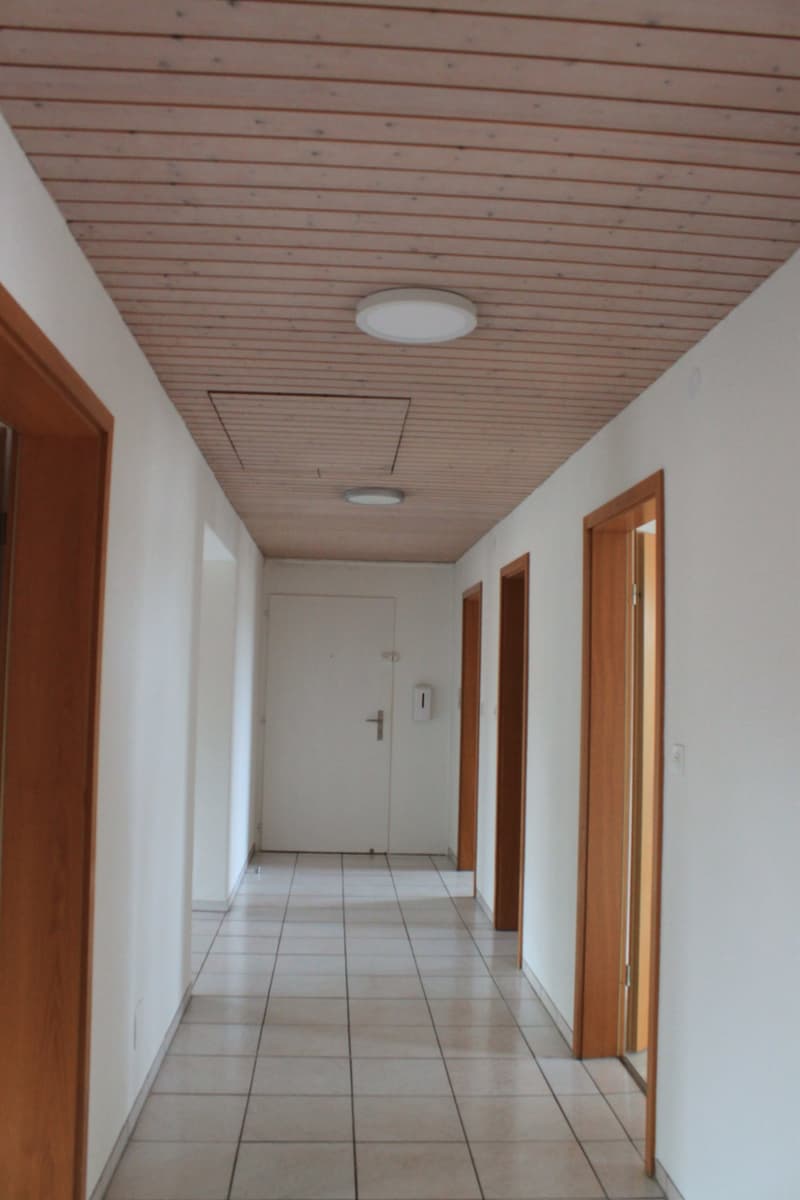 Empty hallway with white tiles, wooden ceiling, and light fixtures.