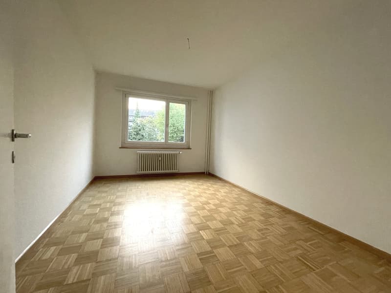 Empty room with wooden parquet flooring, white walls, window with radiator beneath, sunlight coming from the window