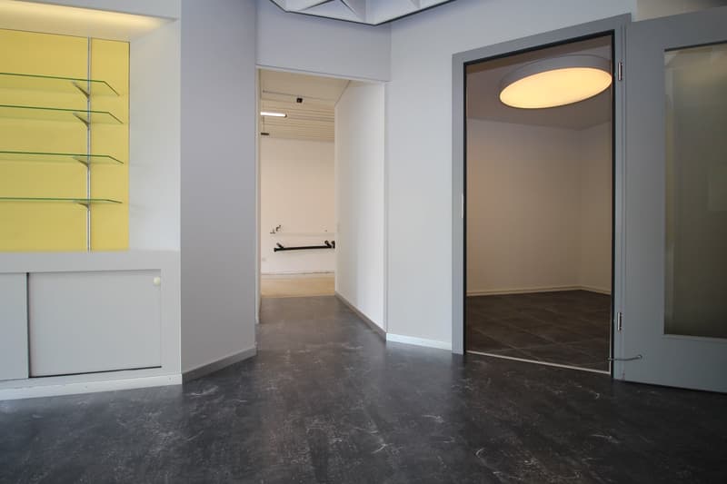 The image shows a modern, minimalist room with gray walls, a large circular light fixture, and a closet or storage space visible in the background. The floor is made of dark, polished concrete. The room has a clean, open layout with minimal furnishings.