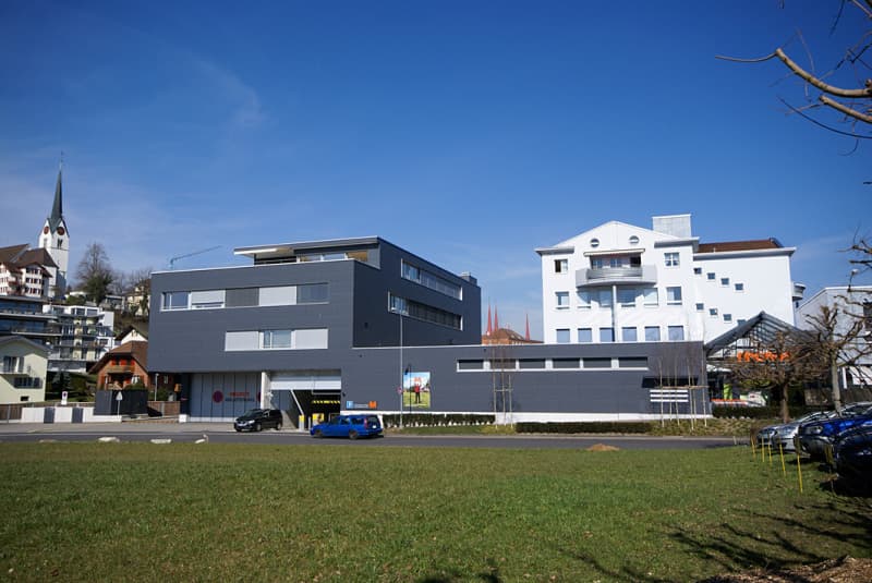 modern grey building with several windows, white building behind, grassy area, cars parked in front