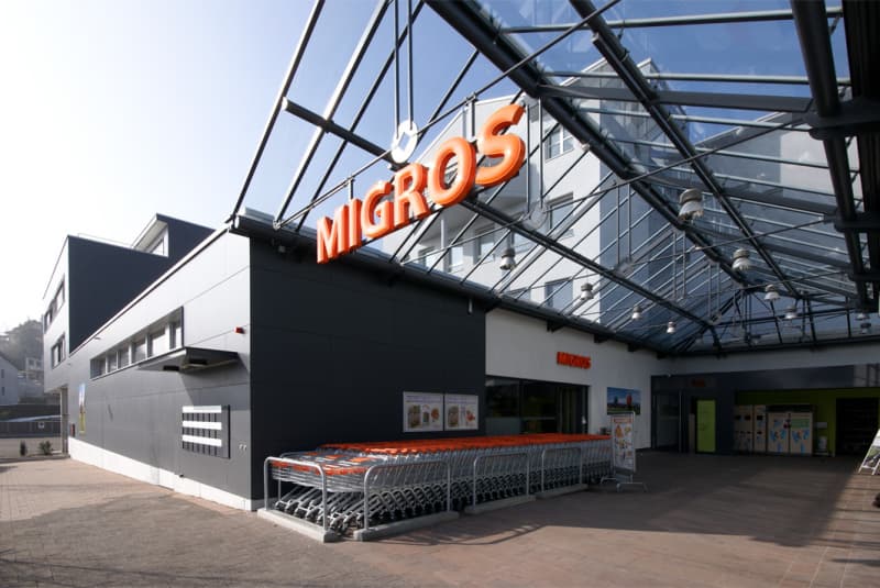 Modern commercial building, large glass windows, black exterior, orange signage for MIGROS, trolley rack outside, large entrance area.