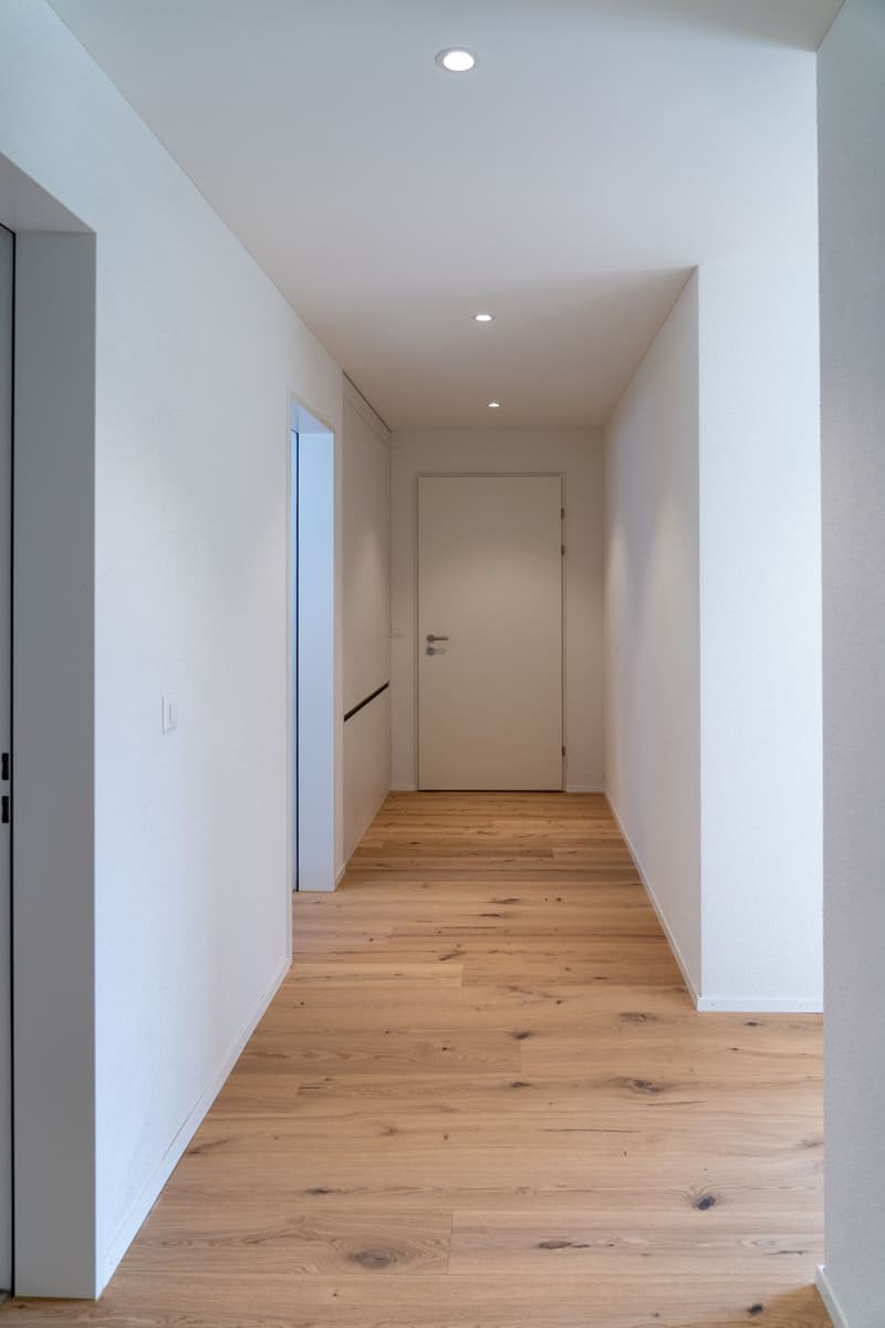 Empty hallway with white walls, wooden floor, recessed lighting, and a white door.