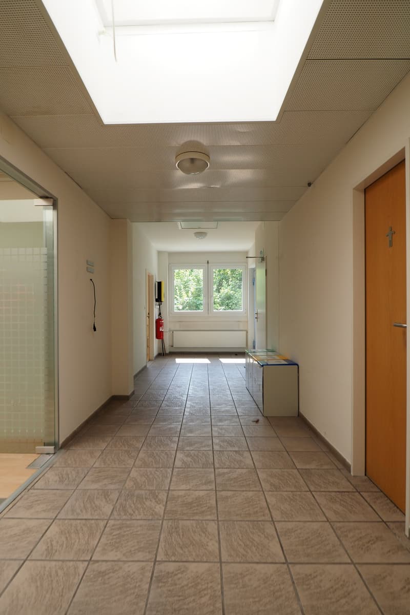 Empty corridor with tiled floor, white walls, two doors, windows, fire extinguisher, electrical outlets, and a fire alarm system