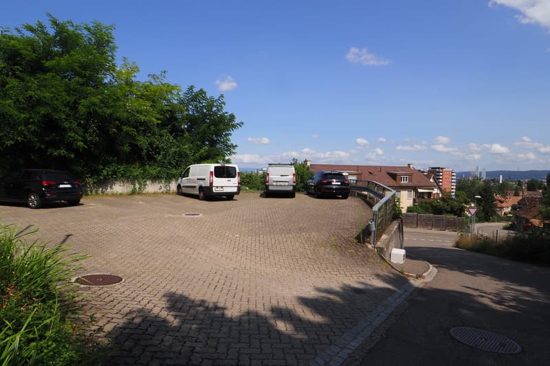 paved parking area with 4 cars parked, surrounded by greenery, with a view of the city