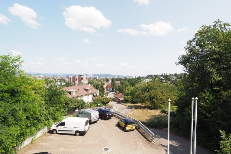 Aerial view of a parking lot with several cars parked, surrounded by trees and greenery, with a distant view of buildings and a city skyline.