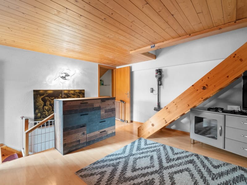Spacious living room with wooden ceiling, white walls, wooden furniture, and a decorative fireplace. The room also features a patterned rug and a piece of artwork on the wall.