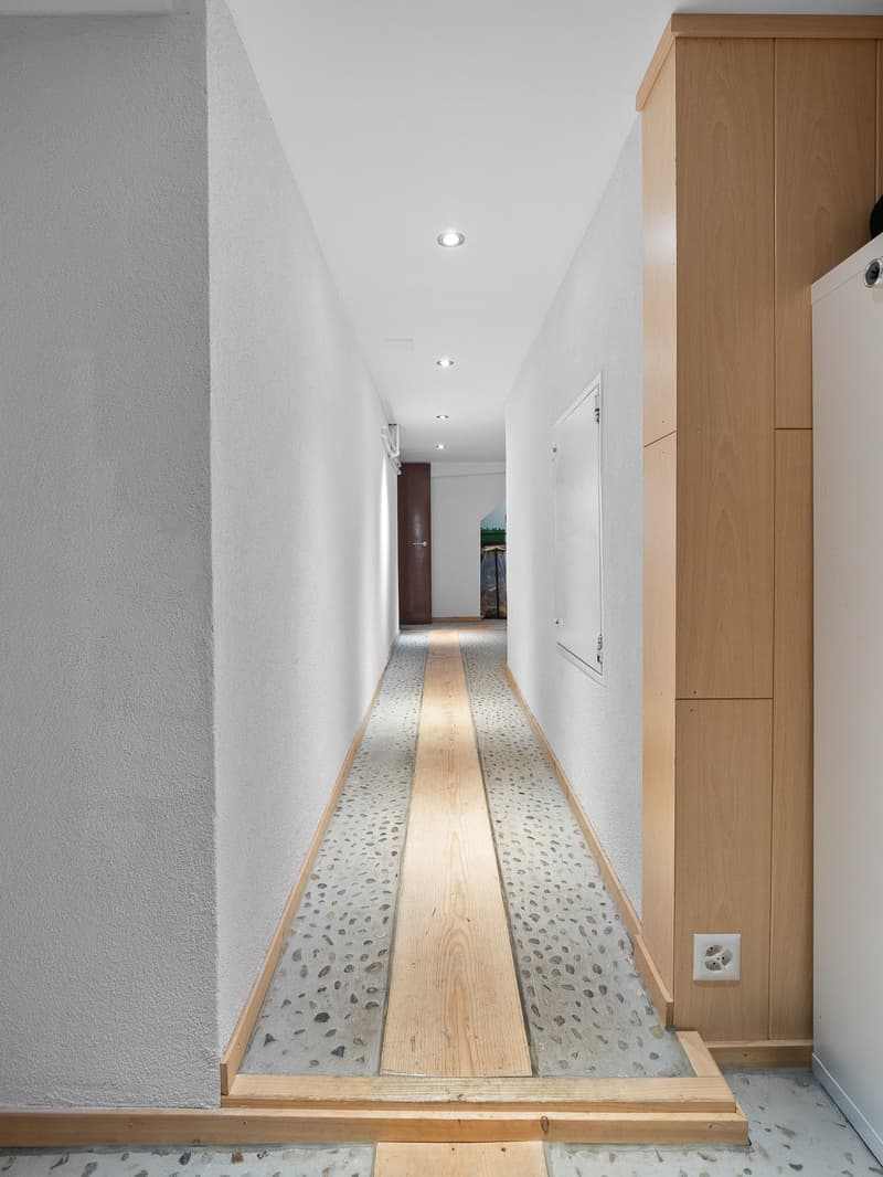 Long, narrow hallway with white walls, wooden floor, and a wooden cabinet along one side. The floor features a mix of wood and pebble-like tiles, creating an interesting textural contrast.