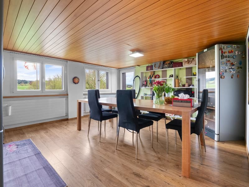 Spacious open-plan living and dining area with wooden ceiling, large windows providing natural light, wooden dining table with black chairs, shelving unit with decorative items, hardwood flooring