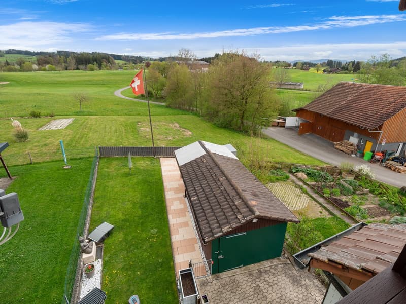 2-story house with a red-tiled roof, surrounded by a grassy field, trees, and a Swiss flag. There is a small shed or outbuilding on the property.